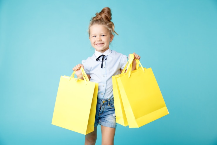 Photo little cute and funny girl holding big bright colored yellow paper bags in the blue studio.