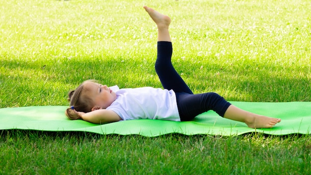 Little cute girl 1-3 in a white t-shirt goes in for sports on a green mat on the grass