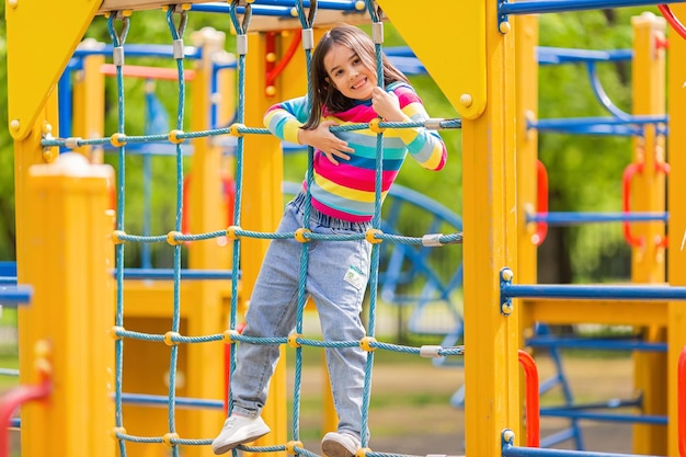 Photo little cute girl 5-6 years old stands on a rope ladder on a childrens ladder on a bright playground