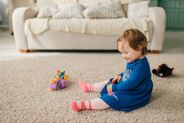 Little cute girl in beautiful dress sits on carpet and plays with her various toys