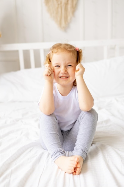 A little cute girl child on a white cotton bed at home is fooling around playing with tails and smiling the child is playing and messing around at home in the morning