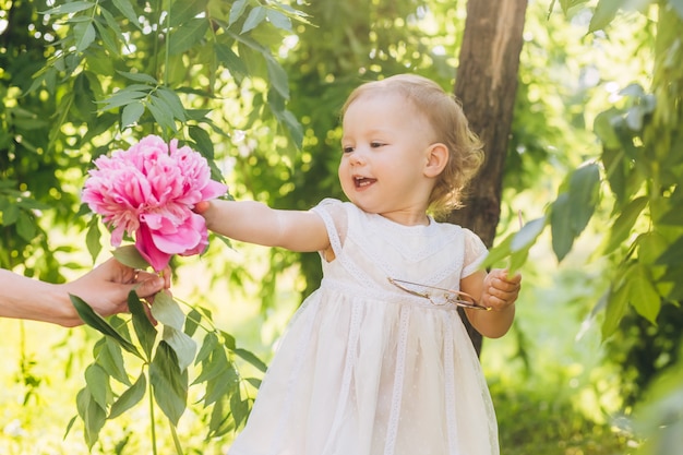 Bambina carina in un abito bianco con una peonia rosa nelle mani nel parco. ritratto di un bambino all'aperto in una giornata di sole estivo. fiori piacevoli della holding della ragazza in sue mani.