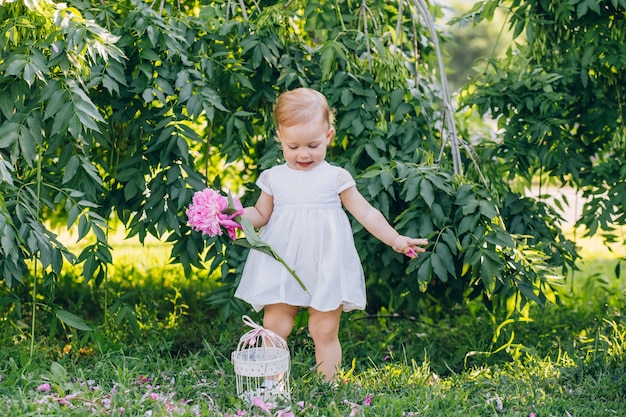 Little cute girl child in a white dress with a pink peony in hands in the park. Portrait of a child outdoors on a sunny summer day. Nice girl holding flowers in her hands.