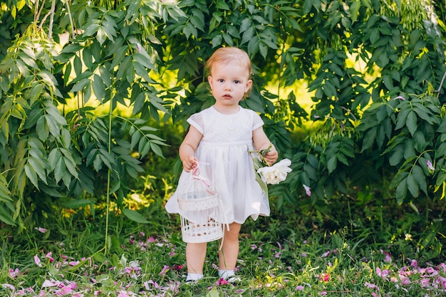 公園で手にピンクの牡丹と白いドレスを着た小さなかわいい女児。日当たりの良い夏の日に屋外の子供の肖像画。手に花を持って素敵な女の子。