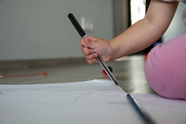 Little cute girl draws on white paper while sitting on the floor. A girl in a white T-shirt and pink pants.