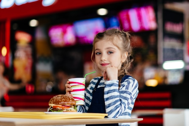 Piccola ragazza carina che mangia un hamburger in un bar, concetto di un pasto veloce per bambini