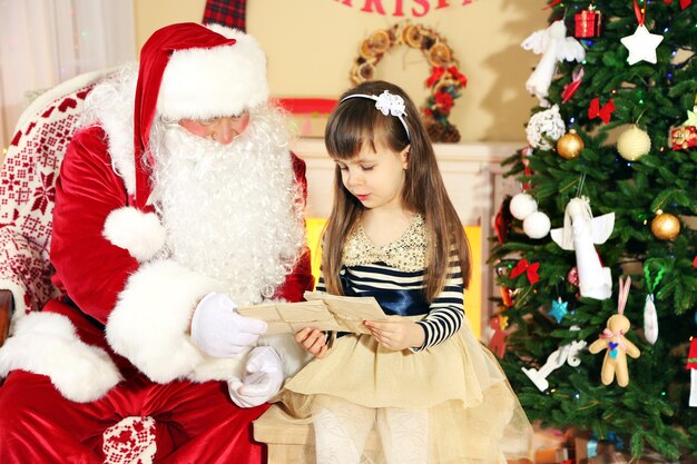 Little cute girl giving letter with wishes to Santa Claus near Christmas tree at home