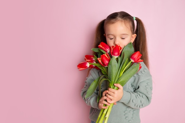 チューリップの花束を持って目を閉じてそれらを嗅ぐ小さなかわいい女の子ピンクの背景。幸せな女性の日。テキスト用のスペース。明るい感情。高品質の写真