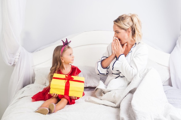 Little cute girl holding present box for young mother looking amazed astonished sitting on bed child wishing happy birthday greeting giving surprise on holiday morning congratulation in bedroom