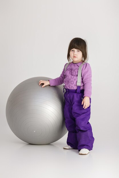 Маленькая милая девочка в фиолетовой одежде с большим мячом для фитнеса на сером фоне.