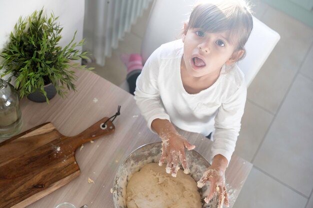 Фото Маленькая симпатичная девочка готовит на кухне, глядя сверху в камеру. веселится, готовя пиццу или тесто для хлеба. детская концепция и весело готовит. скопируйте место для текста слева.