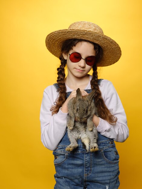 청바지와 밀짚모자, 빨간 선글라스를 쓴 귀여운 소녀가 실제 토끼와 격리된 노란색 배경을 들고 있다