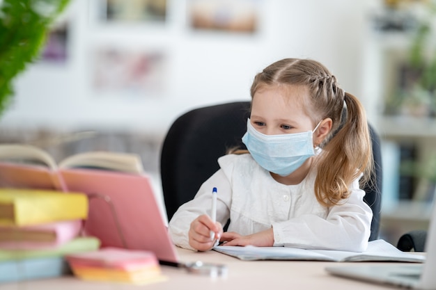 Little cute girl in mask, doing homework, e-learning