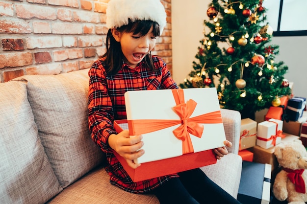 かわいい女の子が驚いた顔でクリスマス ギフト ボックスを開きます。クリスマス プレゼントを見て興奮している陽気な子供。ツリーとテディベアで装飾された部屋で、休日の冬休み、新年が近づいています。