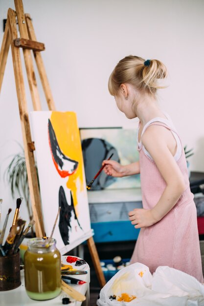 A little cute girl paints a big picture with acrylic at home on an easel