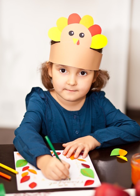 Little cute girl in paper turkey hat writing i am thankful for.\
celebrating thanksgiving day. diy craft art project.