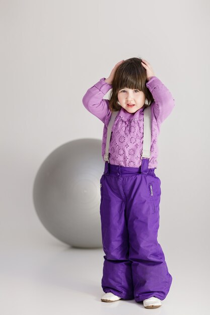 Little cute girl in purple clothes holding his head and large ball for fitness on gray background.