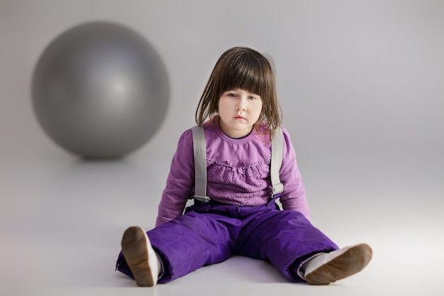 Little cute girl in purple clothes with a large ball for fitness on gray background.