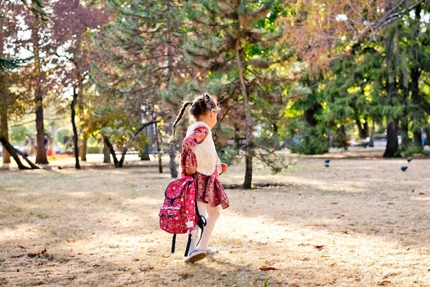 赤いドレスを着た小さなかわいい女の子女子高生がバックパックを持って秋の公園を歩く