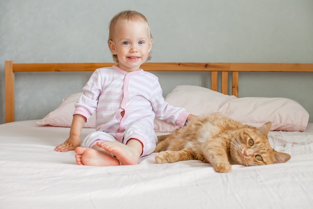 小さなかわいい女の子が太った生姜猫のストロークでベッドに座って、彼と遊ぶ