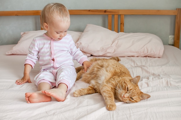 A little cute girl sits on the bed with a fat ginger cat strokes and plays with him