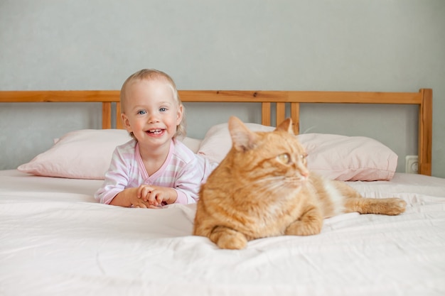 小さなかわいい女の子が太った生姜猫のストロークでベッドに座って、彼と遊ぶ