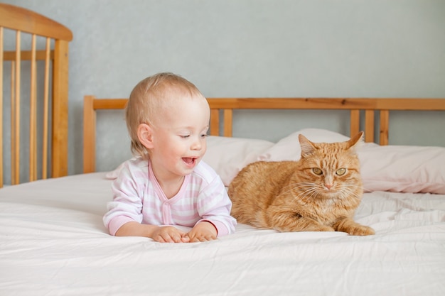 A little cute girl sits on the bed with a fat ginger cat strokes and plays with him