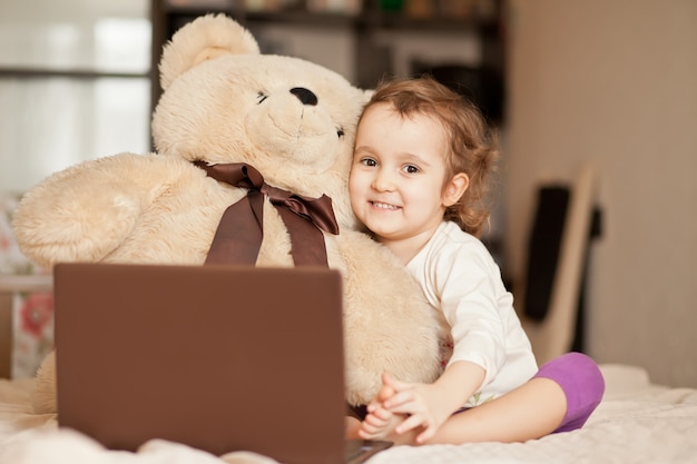 Little cute girl sitting on the bed with big teddy bear and using a digital tablet laptop notebook. Online call friends or parents.