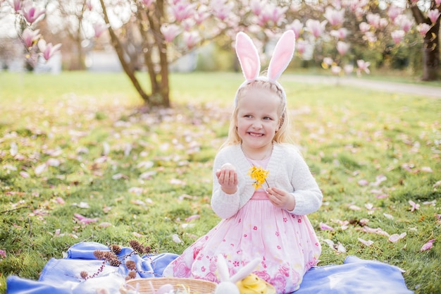 Little cute girl sitting on the grass near the magnolia. A girl dressed as an Easter bunny holds a flower and an egg. Spring.