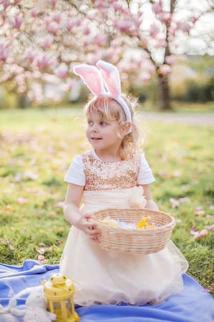 マグノリアの近くの草の上に座っている小さなかわいい女の子。イースターバニーに扮した女の子が花と卵を持っています。春。