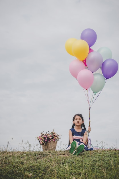 Little cute girl sitting on long green grass outside. Girl holding colorful balloons in hand.