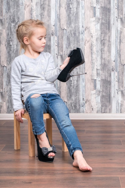Фото Маленькая милая девочка сидит на деревянном стуле и примеряет материнские туфли, изучая их