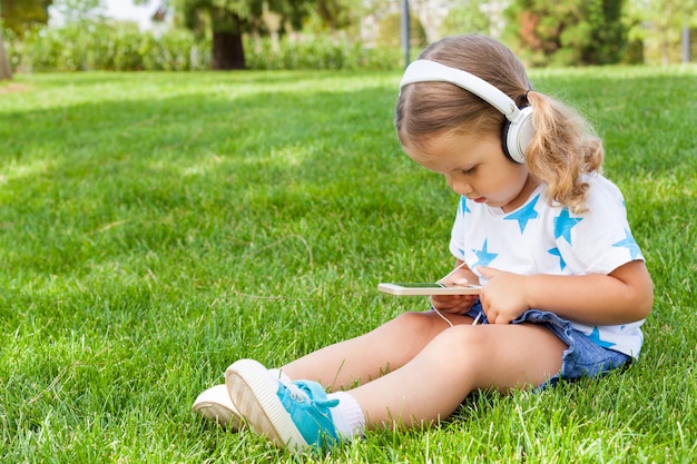 Little cute girl sitting in a Park listening to music in white headphones