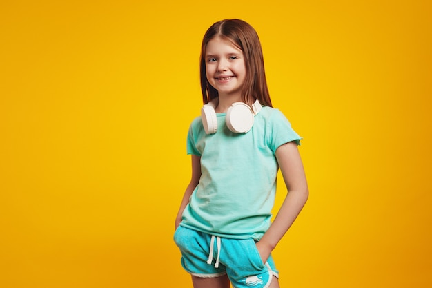 Little cute girl wearing white headphones on neck keeping hands in pockets