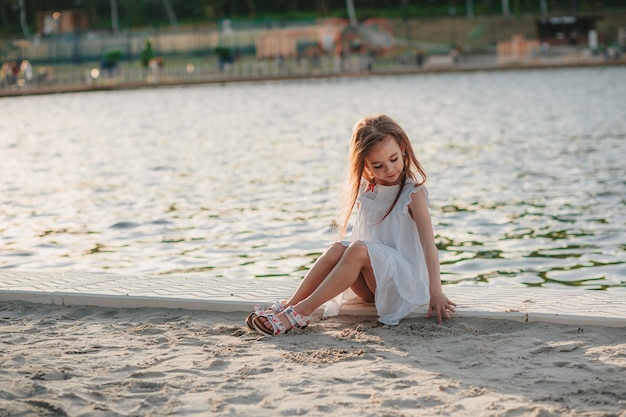 하얀 드레스를 입은 귀여운 소녀가 여름 시간에 해변의 모래 위에 앉아 있다