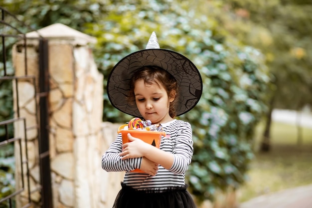 Little cute girl in witch costume holding jack-o-lantern pumpkin bucket with candies and sweets. Kid trick or treating in Halloween holiday.