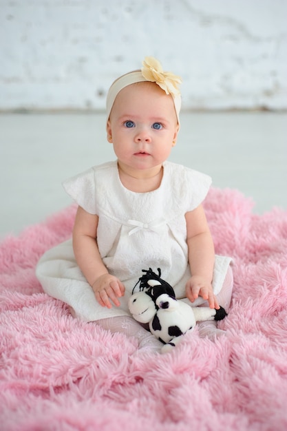 Little cute girl with big blue eyes. The girl is dressed in a white dress. A girl sits on a pink piece of fur against a white brick wall.