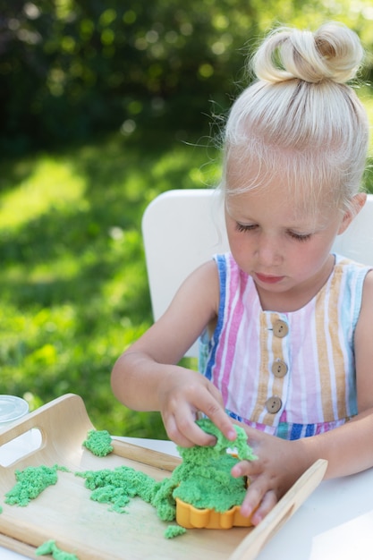 Little cute girl with blonde hair playing with colorful kinetic sand outdoors in the backyard in summer day. Hobby, leisure activity, modeling, crafting. Vertical format.