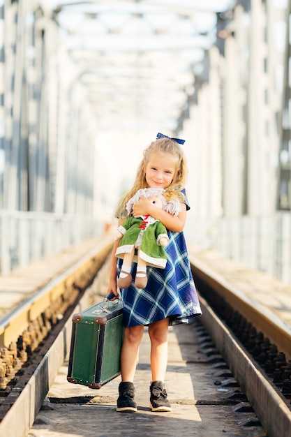 A little cute girl with a large suitcase on a deserted railway platform hugs a doll smiles