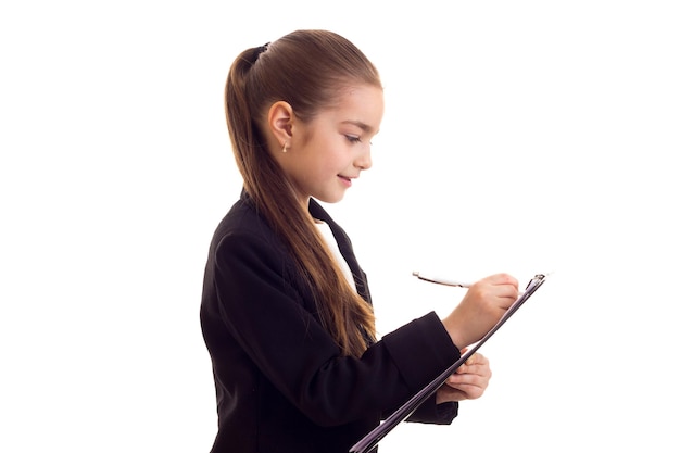 Little cute girl with long brown ponytail in white T-shirt and black jacket holding pen and black folder on white background in studio