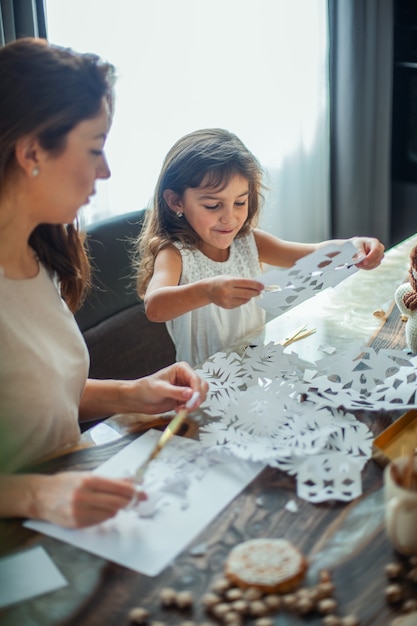 Little cute girl and young beautiful woman cut snowflakes from white paper. Gingerbread and cocoa with marshmallows. The concept of preparation for the New Year and Christmas.