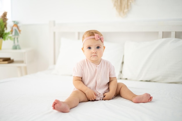 A little cute healthy girl up to a year old in a pink bodysuit made of natural fabric is sitting on a bed on white bed linen in the bedroom looking at the camera the baby is at home