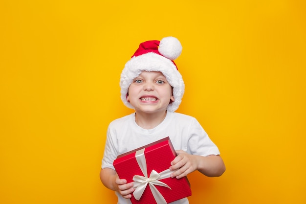 A little cute kid in a white tshirt and a red Santas hat holds the gift box with white ribbon