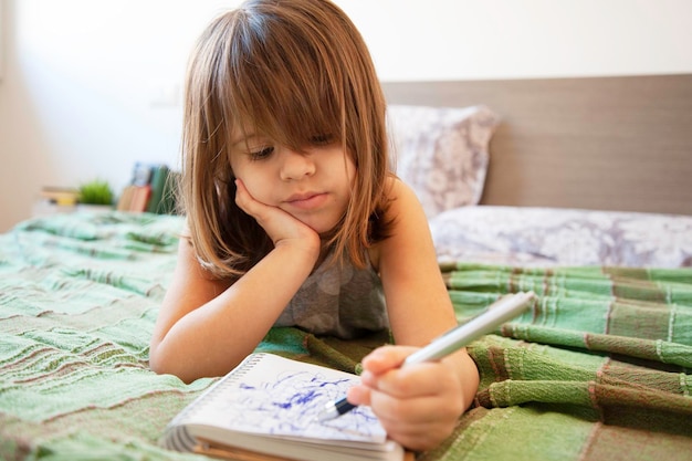 写真 家の寝室のベッドに横たわっている小さなかわいい就学前の女の子彼女は退屈していて、紙のノートに青いペンで走り書きしています子供の頃のコンセプト屋内でのレジャー活動家で退屈する