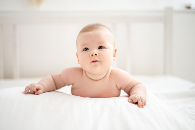 A little cute smiling girl child naked is lying on the bed in the bedroom of the house on white bedding on her stomach looking at the camera smiling happy baby