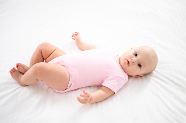 A little cute smiling girl child in a pink bodysuit is lying on the bed in the bedroom of the house on white bedding on her back looking at the camera smiling happy baby