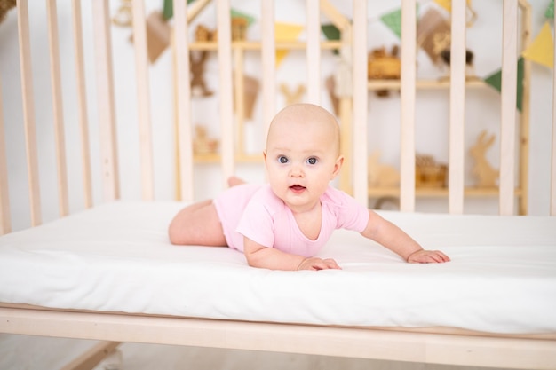 A little cute smiling girl child in a pink bodysuit is lying in a crib at home on white bedding on her stomach looking at the camera smiling happy baby