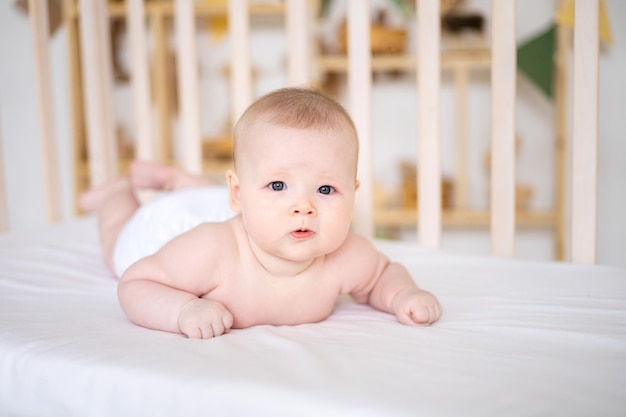 A little cute smiling girl child in white panties is lying in a crib at home on white bedding on her stomach looking at the camera smiling happy baby