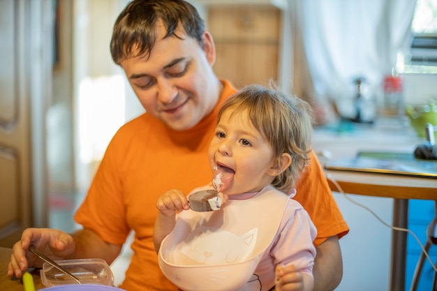 パパの膝の上に座って、ソフトフォーカスのアイスクリームアイスキャンディーを食べる小さなかわいい幼児の女の子。