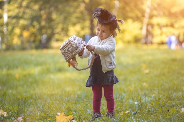 Little cute witch flips a basket and pours autumn leaves in the park.